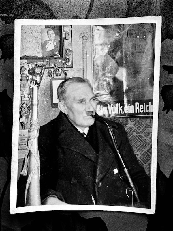 Schwarzweißbild. Ein Türmer sitzt mit einer langen Pfeife im Mund und einem kurzen Horn neben sich in einem Raum. Hinter sich an der Wand ein Hitler-Propaganda-Plakat.
